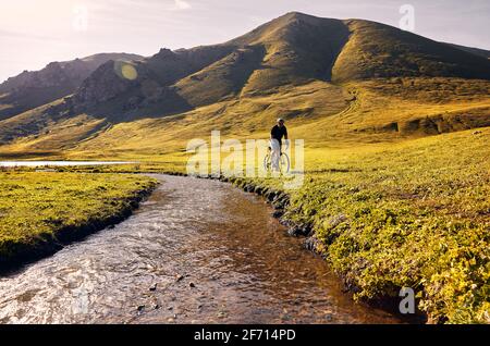 Mann auf Mountainbike-Fahrten in der Nähe des Flusses im grünen Bergtal bei Sonnenaufgang. Freizeit, Reisen und gesundes Lifestyle-Konzept. Stockfoto