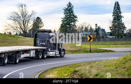 Klassischer schwarzer Langstrecke amerikanischer großer Rig Motorhaube Semi-Truck Traktor mit leerem Flachbett-Sattelauflieger, der den anfährt Runde Autobahneinfahrt zwischen zwei Stockfoto