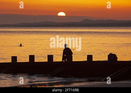 Portobello, Edinburgh, Schottland, UK Wetter. April 2021. Atemberaubender Sonnenaufgang am Ostersonntag am Meer, Temperatur 5 Grad Celsius. Im Bild: Ein junger Mann stellt seine Kamera auf und macht Selfie-Portraits am Strand Groyne am Firth of Forth. Stockfoto