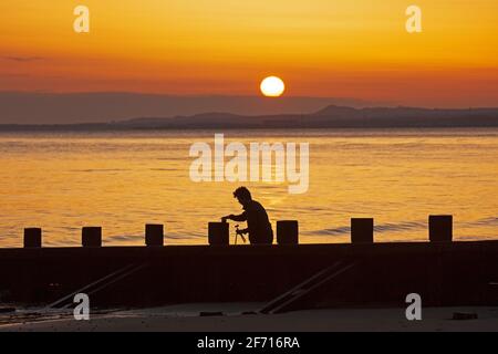 Portobello, Edinburgh, Schottland, UK Wetter. April 2021. Atemberaubender Sonnenaufgang am Ostersonntag am Meer, Temperatur 5 Grad Celsius. Im Bild: Ein junger Mann stellt seine Kamera auf und macht Selfie-Portraits am Strand Groyne am Firth of Forth. Stockfoto