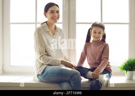 Porträt einer glücklichen jungen Mutter und Tochter, die darauf sitzen Fensterbank zu Hause Stockfoto