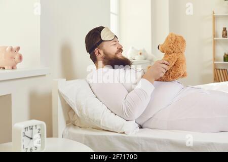 Ein fröhlicher Mann in weißem Schlafanzug und einer Schlafmaske wachte auf und benimmt sich wie ein Kind. Stockfoto