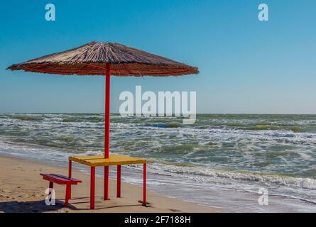 Sommerlandschaft: Tisch, Bänke und Sonnenschirm am Strand. Goldener Sand, stürmisches und winkendes Meer und blauer Himmel. Stockfoto