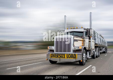 Klassischer dunkler Langstrecken-Sattelzugmaschine mit großer Motorhaube Mit Übergröße Lastschild auf der Vorderseite Transport fixiert mit Schlingen Schnittholz auf dem Flachbett se Stockfoto