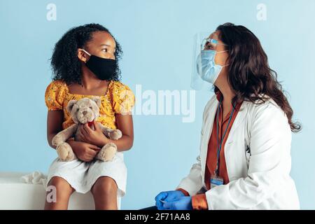Mädchen Besuch Arzt für Check während Pandemie. Ärztin trägt eine schützende Gesichtsmaske und einen Schutzschild, während sie ein kleines Mädchen mit Gesichtsmaske untersucht Stockfoto