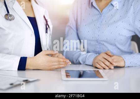 Unbekannte Ärztin zeigt ihrem Patienten eine Beschreibung der Medikamente, während sie zusammen am Schreibtisch in der sonnigen Vitrine in einer Klinik sitzt Stockfoto