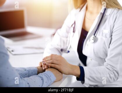 Unbekannte Ärztin hält die Hände ihres Patienten, um den Patienten zu beruhigen, und diskutiert die aktuelle Gesundheitsuntersuchung, während sie zusammen sitzen Stockfoto