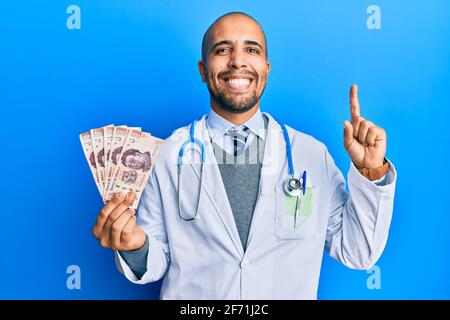 Hispanischer Erwachsener Arzt Mann trägt medizinische Uniform hält 500 mexikanische Pesos lächelnd mit einer Idee oder Frage zeigen Finger mit glücklichem Gesicht, Nummer o Stockfoto