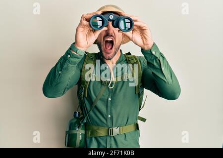 Ein hübscher Mann mit Bart, der einen Explorer-Hut trägt und wütend durch ein Fernglas schaut, frustriert und wütend schreit und wütend schreit und wütend aufschaut Stockfoto