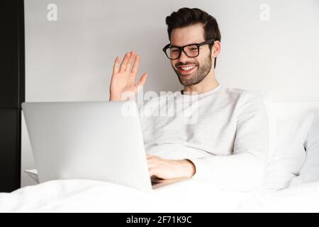 Lächelnder junger Mann winkt mit der Hand und benutzt einen Laptop im Bett Zu Hause Stockfoto
