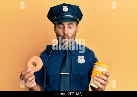 Gutaussehender hispanischer Polizeimann, der Donut isst und Kaffee trinkt und Fisch mit Mund und blinden Augen macht, verrückt und komisch. Stockfoto