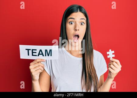 Junge Brünette Frau hält Teampapier und Puzzleteil als Teamarbeit Angst und schockiert mit Überraschung und staunenden Ausdruck, Angst und aufgeregt Gesicht. Stockfoto