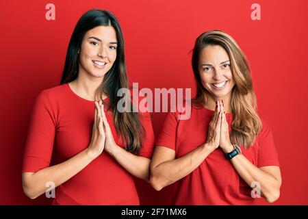 Die hispanische Familie von Mutter und Tochter trägt legere Kleidung Roter Hintergrund beten mit Händen zusammen um Vergebung bitten lächelnd Selbstbewusst Stockfoto