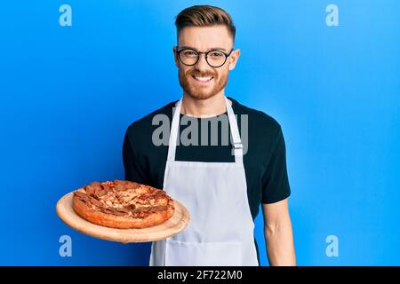 Der junge Rotschopf mit der Kellnerschürze, der Pizza hält, sieht positiv aus Und glücklich stehen und lächeln mit einem selbstbewussten Lächeln zeigen Zähne Stockfoto