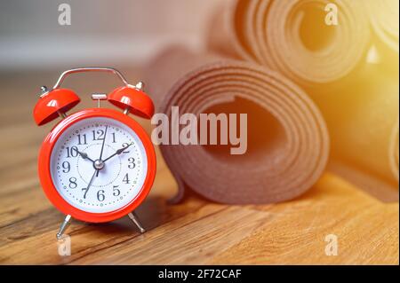 Mehrere gerollte Yoga- oder Fitness-Gummimatten in schwarzer Farbe und roter Wecker auf Holzboden. Sport-Accessoires. Zeit, um sich selbst Konzept zu kümmern. Stockfoto