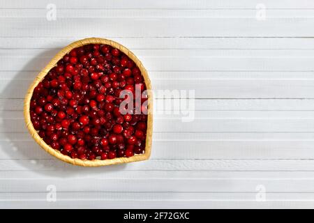 Preiselbeeren in einer Weidenschale in Form eines Herzens. Links schräg auf einem weißen Tisch aus Brettern. Stockfoto