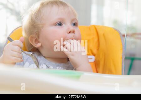 Kleines Kind sitzt auf einem Hochstuhl und isst mit einem Löffel. Nahaufnahme Stockfoto