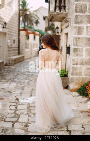 Eine Braut in einem stilvollen Kleid mit offenem Rücken steht vor einem alten Gebäude in einer gemütlichen Perast-Straße, Rückansicht Stockfoto