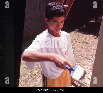 San Antonio., TX USA: Der hispanische Junge malt während einer Freiwilligenarbeit die Außenseite eines Hauses, um einkommensschwachen Bewohnern bei der Verbesserung ihres Wohnhauses zu helfen. Stockfoto