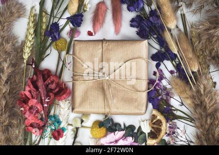 Geschenkbox in umweltfreundliches Bastelpapier verpackt, umgeben von bunten frisch getrockneten Blumen Draufsicht, Frühling, Muttertag, Geburtstag, Grußkarte isoliert Stockfoto