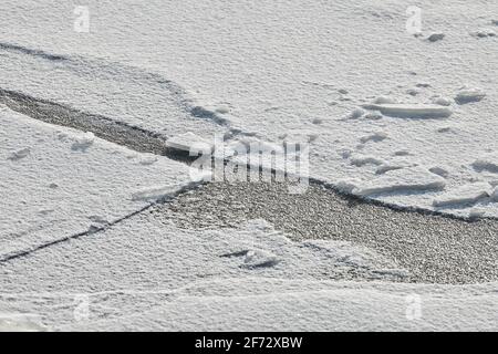 Das Eis des gefrorenen Sees mit weißem Schnee auf der Oberseite ist geknackt. Hintergrund mit Eisstruktur, Nahaufnahme. Stockfoto