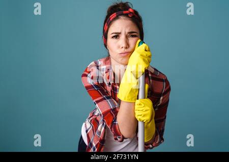 Unglücklich müde junge Frau in Handschuhen posiert mit Mopp isoliert Auf blauem Hintergrund Stockfoto