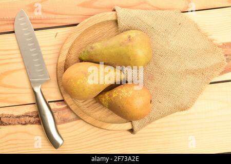 Eine Gruppe von drei reifen Bio-süßen Tasty Conference Birnen auf einem runden Bambustablett mit Jute-Serviette und einem Metallmesser auf einem Tisch aus Holz. Stockfoto