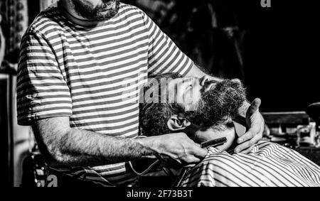 Bärtiger Mann sitzt in einem Sessel in einem Friseurladen, während Friseur seinen Bart mit einem gefährlichen Rasiermesser rasiert. Friseur Rasieren einen bärtigen Mann in einem Stockfoto