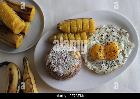 Gedämpfter Hirsemehlkuchen mit einer großzügigen Schicht frischer Kokosnuss dazwischen. Lokal bekannt als Ragi puttu. Serviert mit gedämpftem Kochbananen und Friese Stockfoto