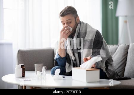 Der kranke Mann, der mit einer Decke bedeckt war, niest in die Nase und nimmt Servietten Stockfoto