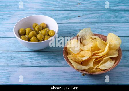 Spanisches Snackkonzept. Gefüllte Oliven in einer weißen Schüssel und Kartoffelchips in einer Holzschüssel auf einem Holztisch in Blautönen Stockfoto