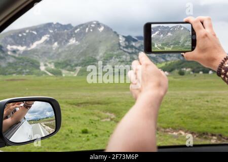 PKW-Mitfahrer, der mit einem Sellphone fotografiert, die Hände durch ein Autofenster streckt, in den Bergen unterwegs ist, der Fokus liegt auf dem Seitenspiegel Stockfoto