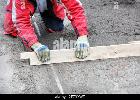 Ein Arbeiter kniet in einem roten Overall das sandige Fundament mit einer Holzebene unter den Pflasterplatten. Kopierbereich, selektiver Fokus. Stockfoto