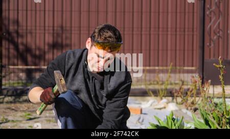 Unschärfe junge Bauarbeiter in gelben Gläsern zerknittert Stirn Entfernen Unregelmäßigkeiten auf Boden Estrich mit Hammer. Männliche Hände in rot, glov Stockfoto