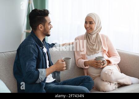 Fröhliches, Schwanger-Muslimisches Paar, Das Sich Zu Hause Ausruhte, Kaffee Trinkt Und Sich Unterhielt Stockfoto
