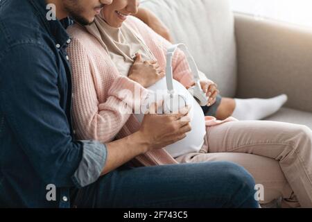 Melodien für Baby in Womb. Erwartungsnahes Paar, das Kopfhörer in der Nähe des trächtigen Bauchs hält Stockfoto