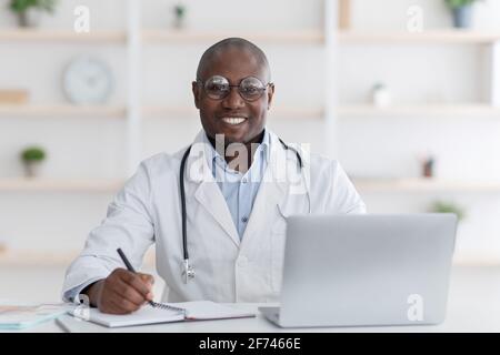 Freundlicher afroamerikanischer Arzt, der in einem modernen Büro mit einem Laptop arbeitet, Notizen macht und seinen Tag plant Stockfoto