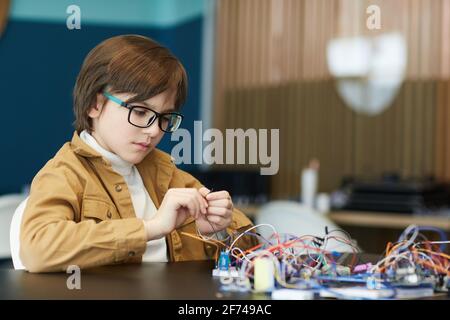 Porträt eines niedlichen Jungen, der eine Brille trägt und mit elektrischen Drähten arbeitet, während er in der Ingenieurklasse Roboter baut und Platz kopiert Stockfoto