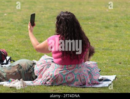 Eine übergewichtige Frau mit dem Rücken zur Kamera sieht aus An ihrem Handy, während sie auf dem Gras saß Ein Park an einem sonnigen Tag Stockfoto