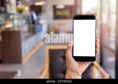Hand Mann hält schwarzes Handy mit weißen Bildschirm mit verwackelte Coffee-Shop Hintergrund. Mockup-Bild der Hand hält Smartphone mit blank weiß Stockfoto