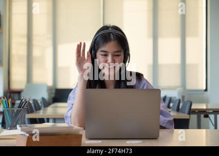 Lächelndes asiatisches Mädchen in Kopfhörern haben Videoanruf entfernte Klasse mit Lehrer mit Laptop, glückliche kleine Kind Welle Gruß mit Tutor, Studie online auf Stockfoto