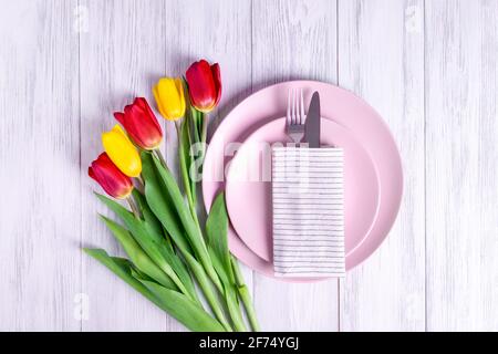 Nahaufnahme eines servierten rosafarbenen Tellers, Besteck in einer Serviette und ein Strauß Tulpen auf einer hellen Holzoberfläche. Konzept der Frühlings-Gratulation. Selektiver Fokus Stockfoto