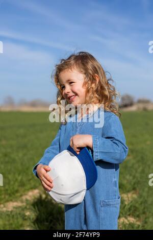 Glückliches blondes Mädchen hält Kappe, während sie wegschaut, gegen sie zu stehen Blauer Himmel im Sommer auf der Wiese Stockfoto