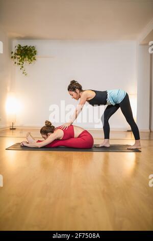 Seitenansicht der weiblichen Ausbilderin, die die Frau beim Yoga-Training auf einer Sportmatte im Studio beim Sitzen nach vorne unterstützt Stockfoto