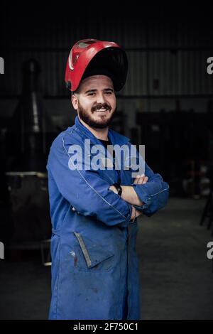 Glücklicher bärtiger Arbeiter mit schützendem Hardhut und blauem Overall In der Garage stehend und mit gekreuzten Armen auf die Kamera schauend Stockfoto