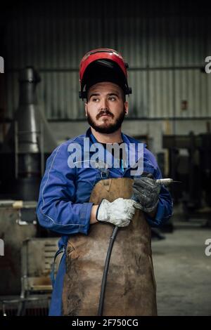 Mechaniker in Schutzhelm und Schürze auf blauem Overall stehend Mit Schweißschlauch in Lichtwerkstatt in der Nähe von Metallkonstruktionen Stockfoto