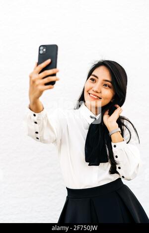 Asiatische Frau mit langen dunklen Haaren, die sich selbst porträtieren Mobiltelefon steht auf weißem Hintergrund Stockfoto
