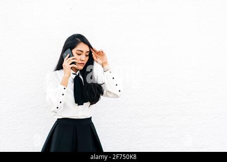 Teen ethnisch asiatische weiblich in formellen Outfit stehen in der Nähe von weiß Pinnwand auf dem Mobiltelefon sprechen Stockfoto