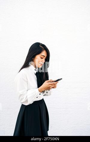 Seitenansicht von Teenagers ethnischer Frau im formellen Outfit stehend In der Nähe einer weißen Wand und mit einem Mobiltelefon Stockfoto