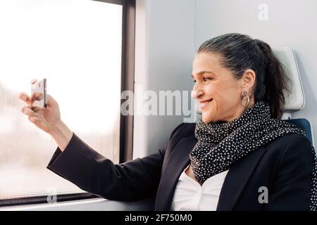 Seitenansicht einer positiven Frau mittleren Alters mit Schals Bild auf dem Mobiltelefon, während Sie auf dem Beifahrersitz in der Nähe des Fensters sitzen Im Wagen während der Fahrt Stockfoto